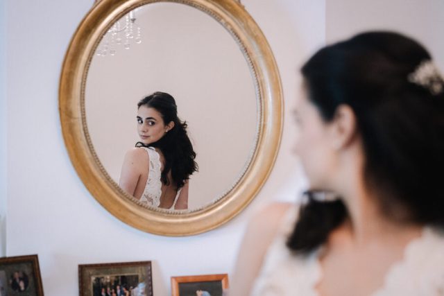 Reflet de la mariée dans un miroir ovale ajustant sa coiffure lors de ses préparatifs.