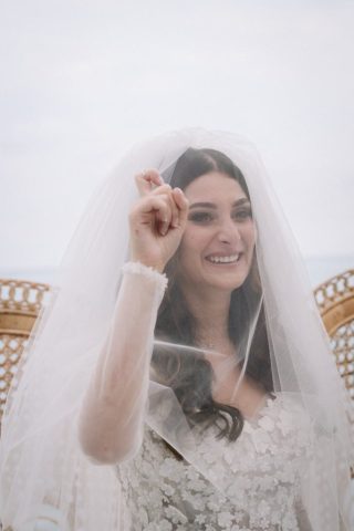 La mariée, sous son voile, montre fièrement son alliance à ses invités en souriant.