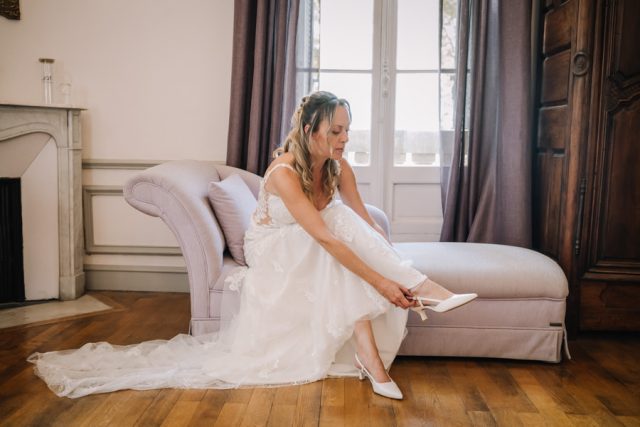 La mariée enfile ses chaussures de mariage assise dans un salon élégant du domaine.