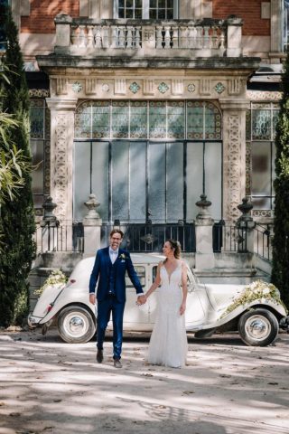 Le couple marche vers une voiture de collection blanche garée devant la verrière du château.