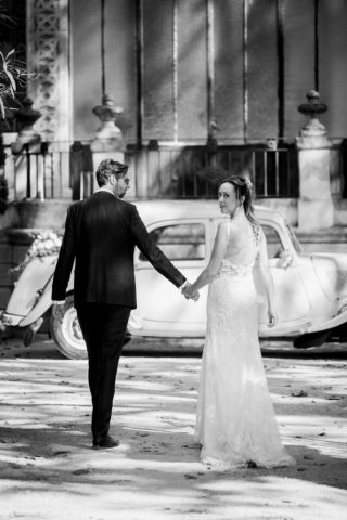 Photographie en noir et blanc des mariés se tenant la main devant le véhicule ancien.
