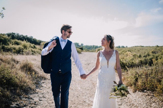 Le couple de mariés marche main dans la main avec complicité sur un chemin en terre.