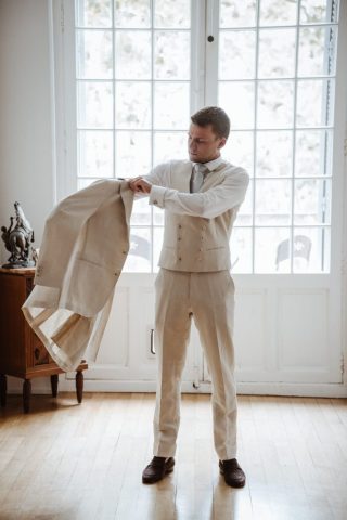 Le marié enfile sa veste de costume beige clair de manière dynamique.
