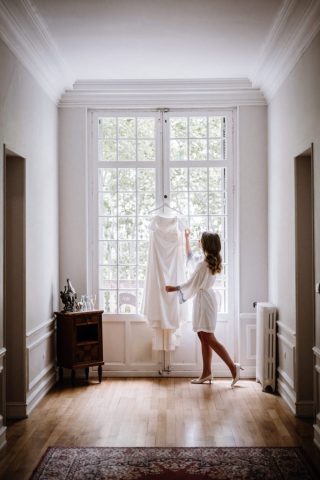 La mariée admire sa robe de mariée suspendue devant une grande fenêtre lumineuse au domaine.