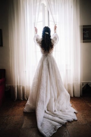 La mariée de dos face à la lumière, dévoilant la longue traîne et la dentelle de sa robe de mariée.