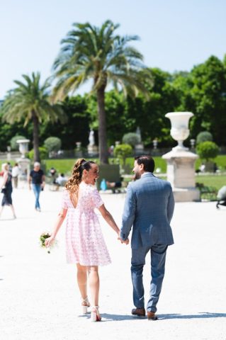 Les mariés s'éloignent de dos dans les jardins parisiens, la mariée portant une robe courte élégante.